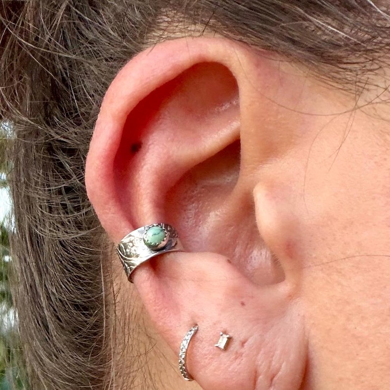May include: Close-up of an ear adorned with a silver ear cuff featuring a small, round, turquoise stone. A small, silver hoop earring and a tiny, square-cut stud earring are also visible. The ear cuff has a detailed, floral design.