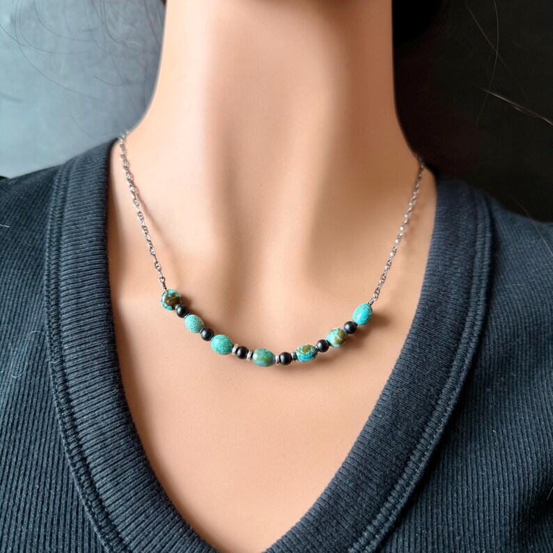 May include: A silver chain necklace with turquoise and black beads. The necklace features a series of round turquoise beads interspersed with smaller black beads. The necklace is worn on a person wearing a black shirt.