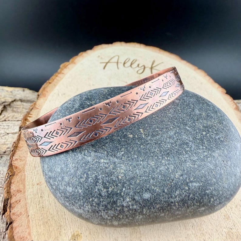 May include: A copper cuff bracelet featuring a repeating geometric pattern of etched chevrons, diamonds, and horizontal lines. The metal has a warm, reddish-brown tone with dark oxidized accents within the engraved design details.