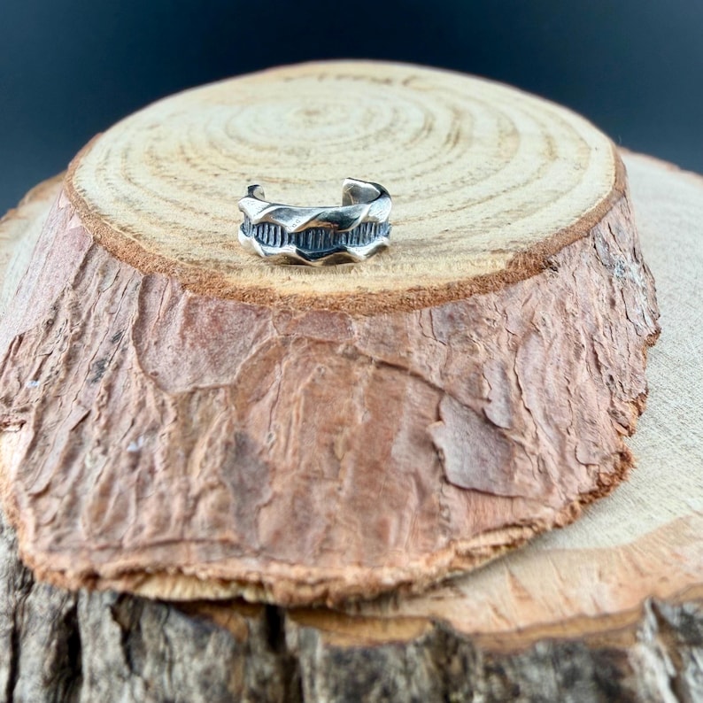 May include: A silver-colored ear cuff with a textured, wave-like design. The cuff is open-ended and wraps around the ear. The jewelry is displayed on a rustic, wooden surface with a natural bark edge.