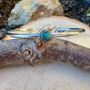 May include: A silver cuff bracelet with a round turquoise stone set in the center. The turquoise stone has a mottled pattern of blue and green. The bracelet is displayed on a piece of wood.