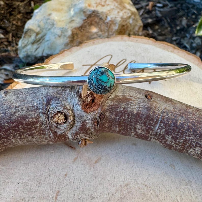 May include: A silver cuff bracelet with a round turquoise stone set in the center. The turquoise stone has a mottled pattern of blue and green. The bracelet is displayed on a piece of wood.