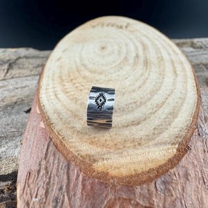 May include: A silver ring with a textured surface and a geometric diamond design. The ring is displayed on a wooden surface, showcasing its handcrafted details and unique style. This piece is perfect for adding a touch of Southwestern flair to any outfit.