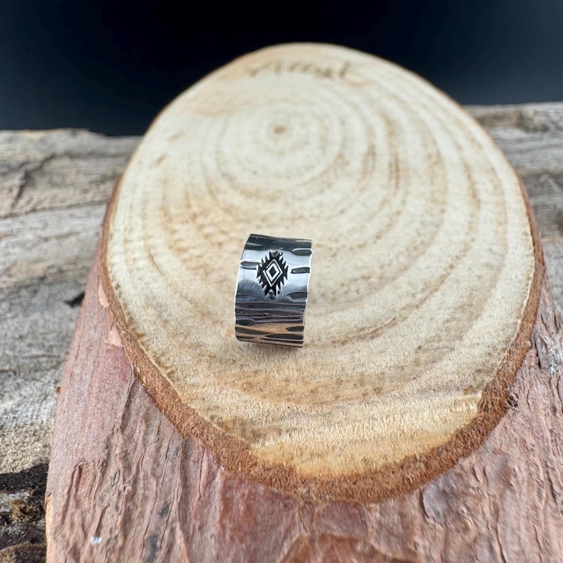 May include: A silver ring with a textured surface and a geometric diamond design. The ring is displayed on a wooden surface, showcasing its handcrafted details and unique style. This piece is perfect for adding a touch of Southwestern flair to any outfit.
