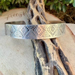 May include: A silver-toned cuff bracelet featuring a repeating geometric pattern of etched diamonds and triangles. The metal band has a polished finish and an open design, showcasing intricate, symmetrical engravings across its entire surface.