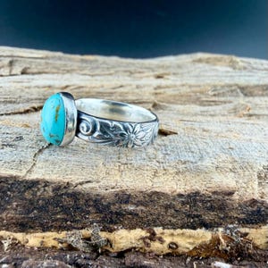 May include: A silver ring with a large, oval turquoise stone. The band is intricately carved with floral and scroll designs. The ring is displayed on a textured wooden surface, with a dark background.