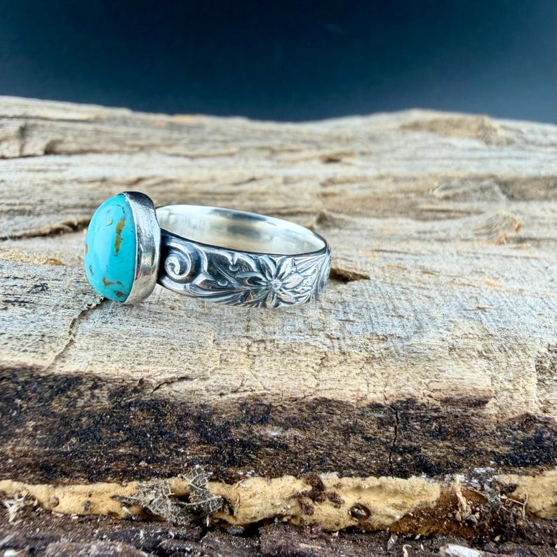 May include: A silver ring with a large, oval turquoise stone. The band is intricately carved with floral and scroll designs. The ring is displayed on a textured wooden surface, with a dark background.