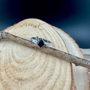 May include: A silver ring with a black, diamond-shaped stone. The ring is set on a wooden branch against a blurred background. The ring's band has a decorative, swirling design.