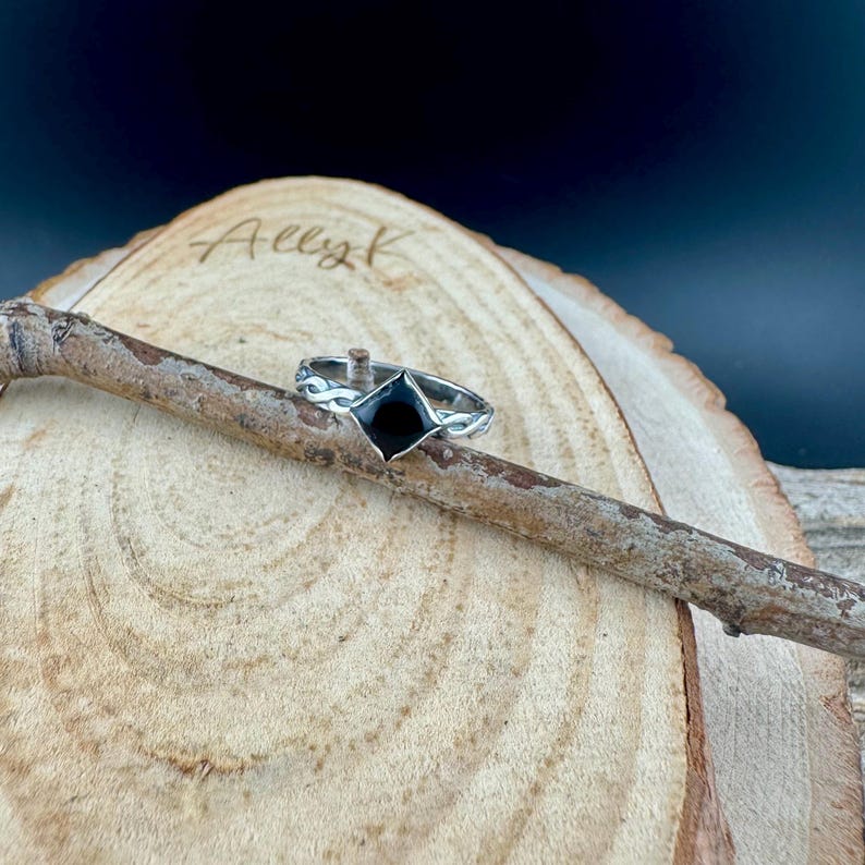 May include: A silver ring with a black, diamond-shaped stone. The ring is set on a wooden branch against a blurred background. The ring's band has a decorative, swirling design.