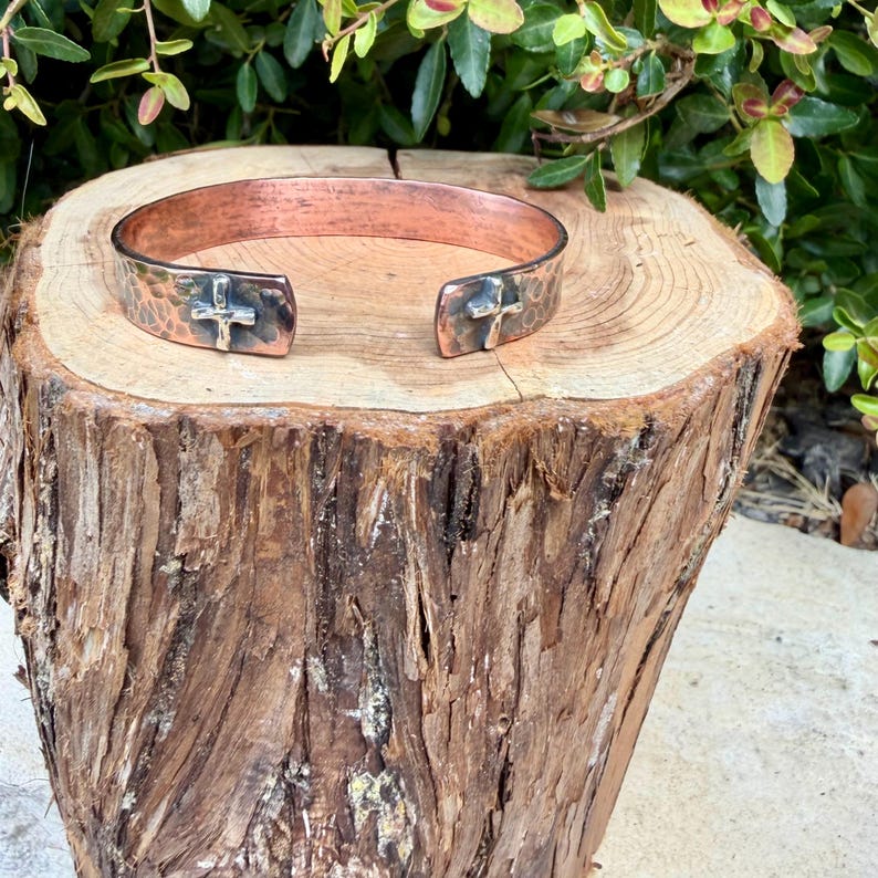 May include: A copper cuff bracelet featuring a hammered texture and a dark, oxidized finish. The open-ended design includes two small, raised silver-toned cross emblems positioned at each terminal end of the metal band.