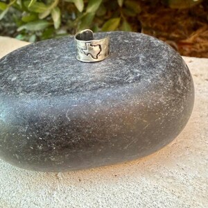 May include: A silver-toned metal ring featuring a cutout silhouette of the state of Texas. The band has a wide, open-ended design with a smooth, polished finish. The ring is positioned on a dark, smooth, rounded stone surface.