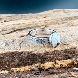 May include: A silver ring with an oval, off-white stone. The band features an engraved wave pattern. The ring is displayed on a textured, wooden surface.