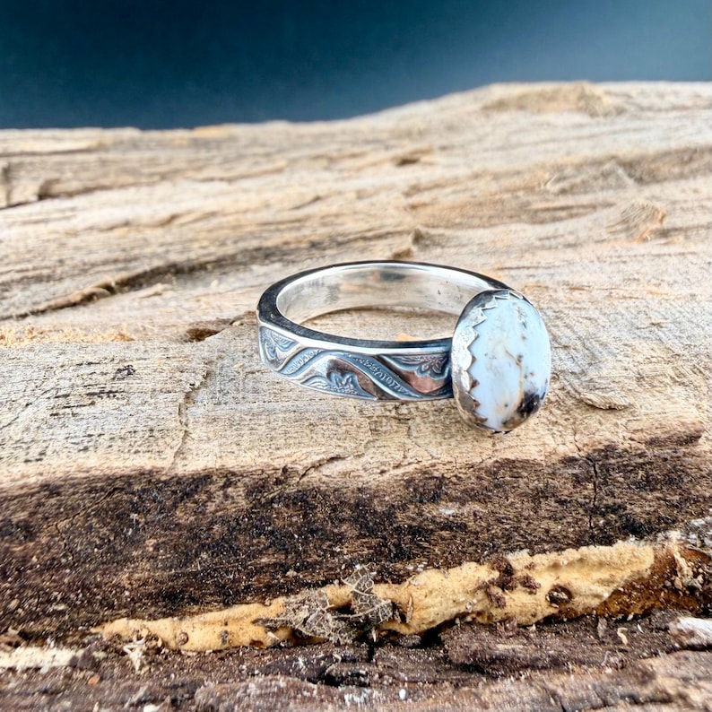 May include: A silver ring with an oval, off-white stone. The band features an engraved wave pattern. The ring is displayed on a textured, wooden surface.