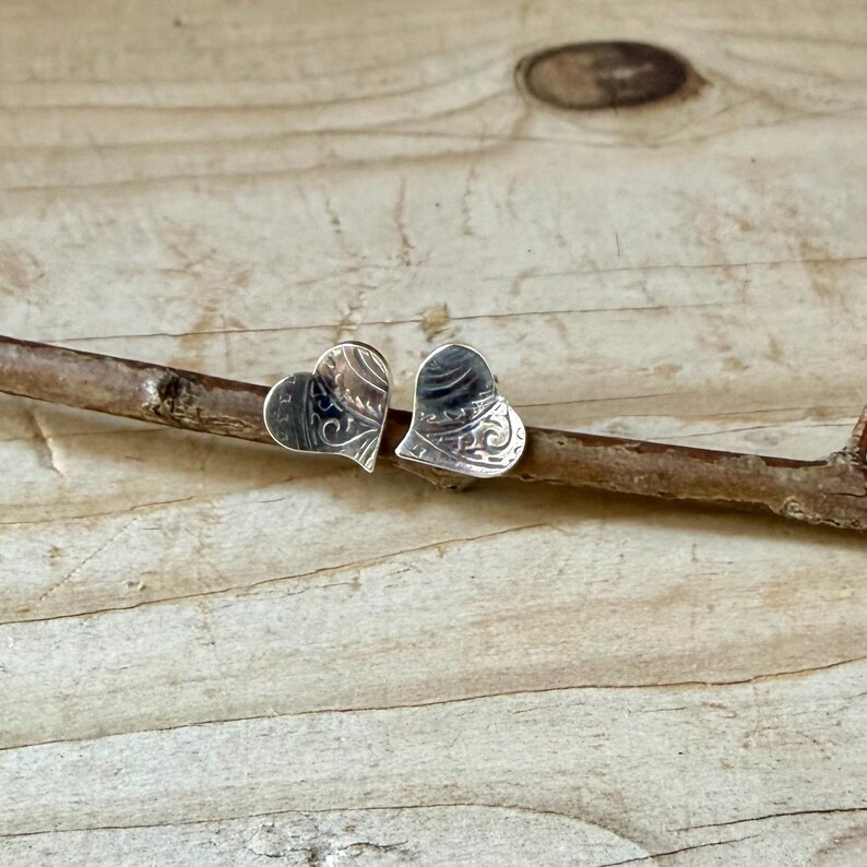 May include: A pair of silver heart-shaped stud earrings featuring an intricate, engraved swirling pattern on the surface. The earrings are positioned side-by-side, resting on a thin, textured brown twig.