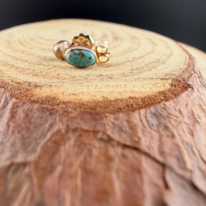 May include: A gold-colored earring with a turquoise stone. The earring is oval-shaped and set in a gold bezel. The earring is on a wooden surface with a dark background.