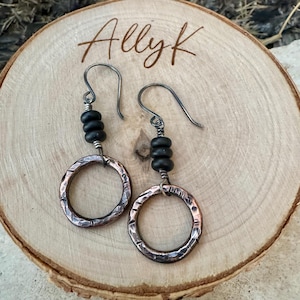 May include: A pair of handcrafted earrings featuring hammered copper hoops and black beads. The earrings have silver-toned ear wires and are displayed on a wooden surface. The name "Allyk" is written on the wood.