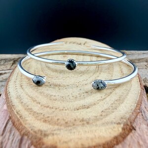 May include: Two silver-toned open cuff bracelets with oval, speckled black and gray stones. The bracelets are displayed on a wooden surface, with a dark background. The bracelets have a simple, elegant design.