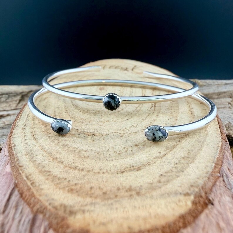 May include: Two silver-toned open cuff bracelets with oval, speckled black and gray stones. The bracelets are displayed on a wooden surface, with a dark background. The bracelets have a simple, elegant design.