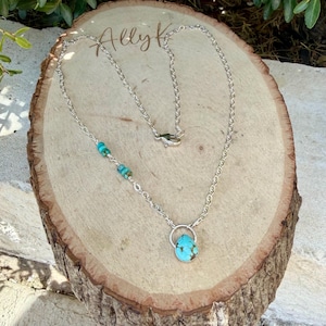 May include: A silver chain necklace featuring a teardrop-shaped turquoise pendant suspended from a circular silver ring. The chain includes three small turquoise beads on one side and a lobster claw clasp closure at the back of the necklace.