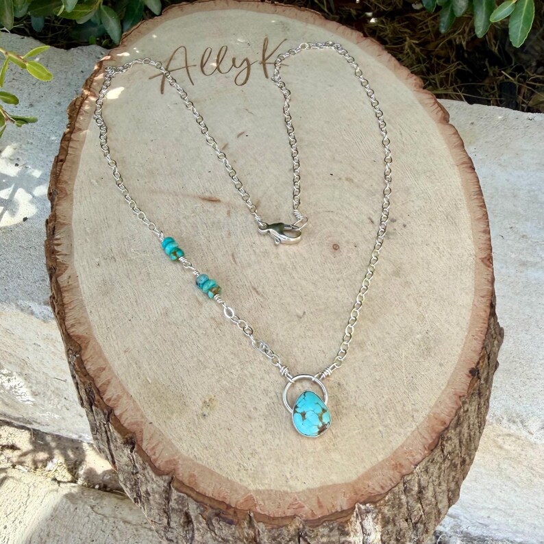 May include: A silver chain necklace featuring a teardrop-shaped turquoise pendant suspended from a circular silver ring. The chain includes three small turquoise beads on one side and a lobster claw clasp closure at the back of the necklace.