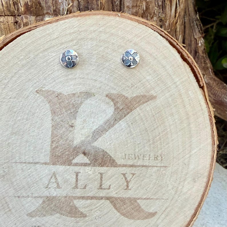 May include: A pair of small, round, silver-toned stud earrings featuring a stamped cross design. The earrings are displayed on a circular wooden slice engraved with the text K ALLY JEWELRY in a large, stylized font across the center.