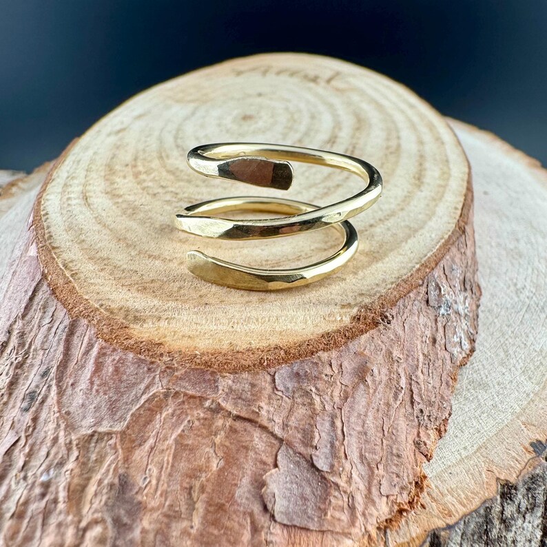 May include: A gold-colored spiral ring with a hammered texture. The ring is displayed on a wooden surface, showcasing its unique design. The ring's design features a wrap-around style, adding a modern touch to the jewelry.