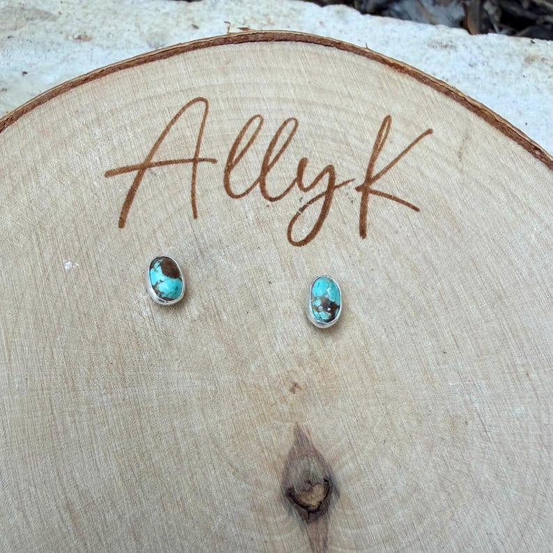 May include: A pair of oval turquoise stud earrings with a silver bezel, displayed on a wooden surface. The earrings have a brown matrix within the turquoise. The name "Allyk" is written in brown script on the wood.