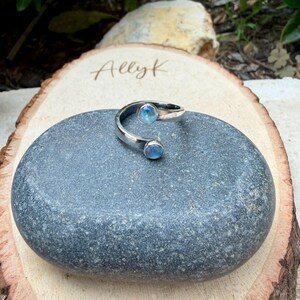 May include: A silver ring featuring two light blue, round gemstones. The ring has a curved design, with a gemstone at each end. It is displayed on a grey stone, with a wooden background.