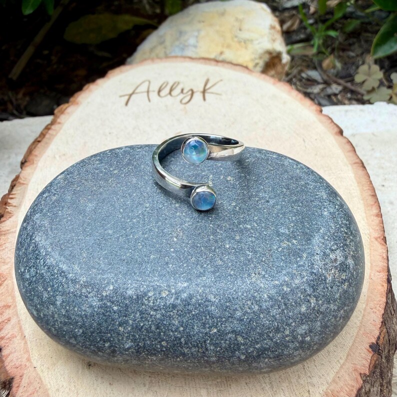 May include: A silver ring featuring two light blue, round gemstones. The ring has a curved design, with a gemstone at each end. It is displayed on a grey stone, with a wooden background.