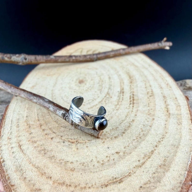 May include: A silver-colored ear cuff with an engraved design and a dark brown gemstone. The cuff is set against a wooden background with a natural branch, showcasing the jewelry's craftsmanship and earthy aesthetic.