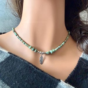 May include: A turquoise beaded necklace with a silver feather pendant. The necklace features small, irregular turquoise beads and a delicate silver feather charm. The necklace is displayed on a person wearing a black and gray plaid shirt.