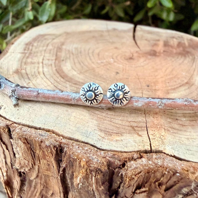 May include: A pair of circular silver stud earrings featuring a raised sun design with radiating rays around a central dot. The earrings are resting on a thin, light brown twig that is positioned horizontally across a round wooden log slice.