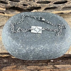 May include: A silver chain bracelet with a rectangular pendant featuring a cutout of the state of Texas. The bracelet is displayed on a grey stone surface with a wooden background.