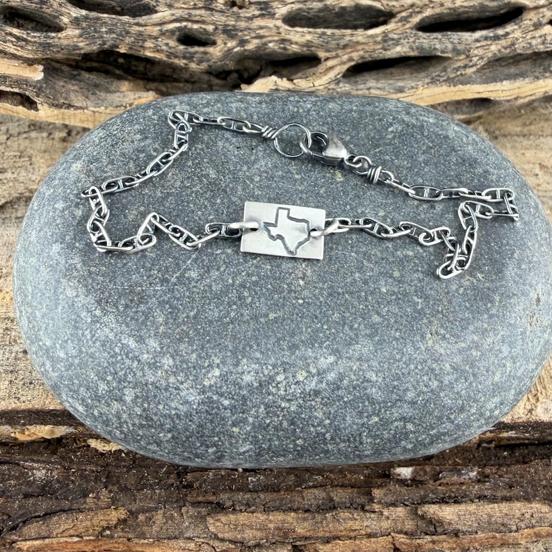May include: A silver chain bracelet with a rectangular pendant featuring a cutout of the state of Texas. The bracelet is displayed on a grey stone surface with a wooden background.