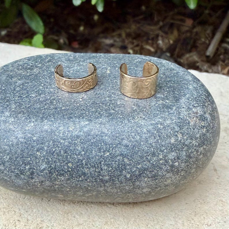 May include: Two gold-colored ear cuffs with a floral design. The ear cuffs are made of a shiny metal and are placed on a gray stone. The cuffs have a curved shape and are open at the bottom.