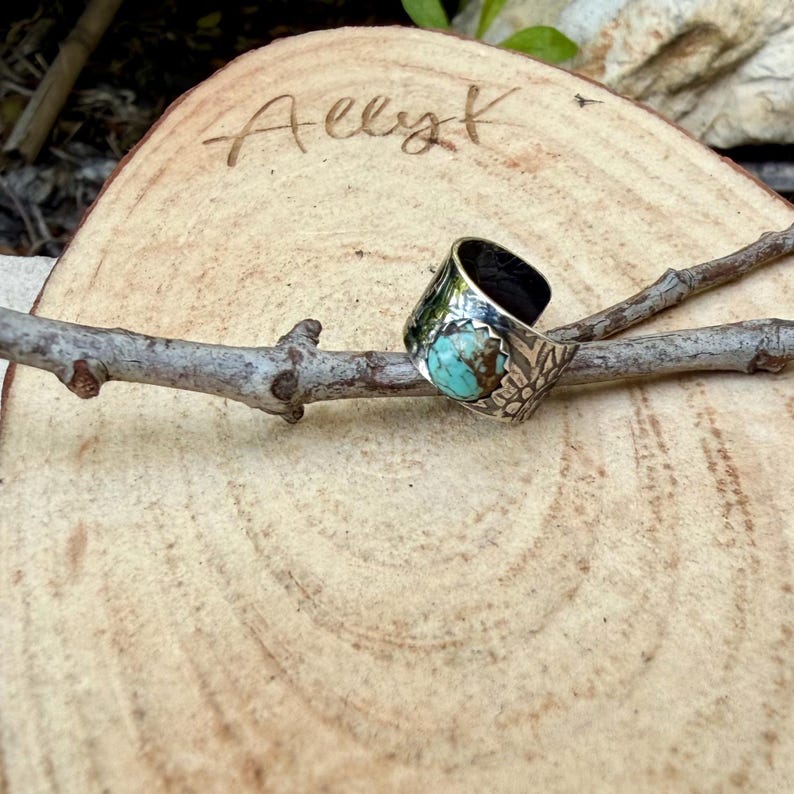 May include: A silver ring with a turquoise stone and floral design. The ring is set against a wooden background with the handwritten word "Allyk" carved into it. The ring is on a small branch.