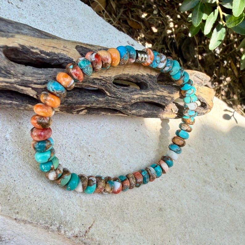 May include: A beaded bracelet featuring round, flat-disc shaped stones in vibrant shades of turquoise, orange, and white with natural veining. The beads are strung together in a circular pattern and include a single small, metallic silver-toned spacer.