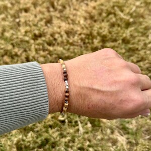 以下が含まれることがあります： 茶色、ベージュ、シルバー色のビーズを組み合わせたビーズブレスレット。 ブレスレットは細くてしなやかなコードに通されています。 ビーズは様々なサイズと形状で、自然でアースカラーの美しさを演出しています。 ブレスレットは手首に装着されています。