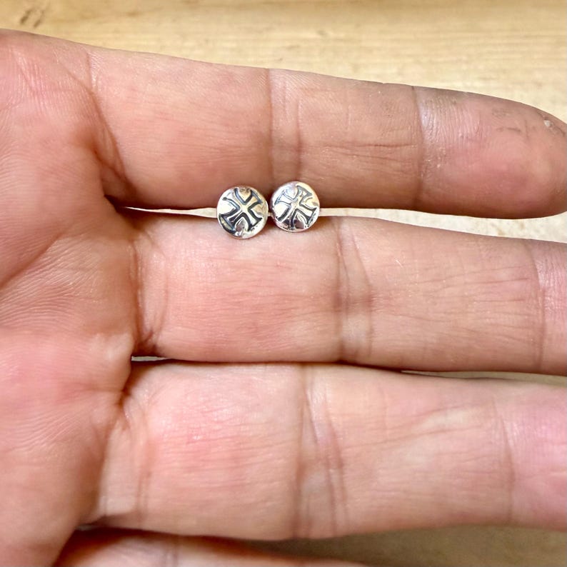 May include: A pair of small, circular silver stud earrings. Each earring features a polished, metallic surface with a recessed, cross-shaped design stamped into the center. The studs have a smooth, rounded edge and a reflective, bright silver finish.