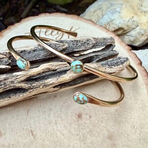 May include: Two gold-colored metal cuff bracelets with turquoise-colored stones. The bracelets are curved and rest on a piece of driftwood and a wooden surface. The turquoise stones have brown marbling.