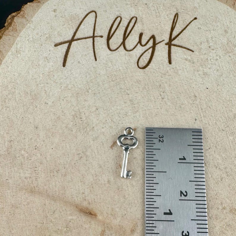 May include: A small, silver-colored key charm next to a ruler. The key is approximately 1 inch long. The background is a light brown, wooden surface with the handwritten text "Allyk" in brown cursive.
