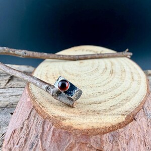 May include: A silver ring with a rectangular setting and a round, deep red gemstone. The ring is set against a natural wood background, with a dark backdrop. The ring's band has a textured appearance.