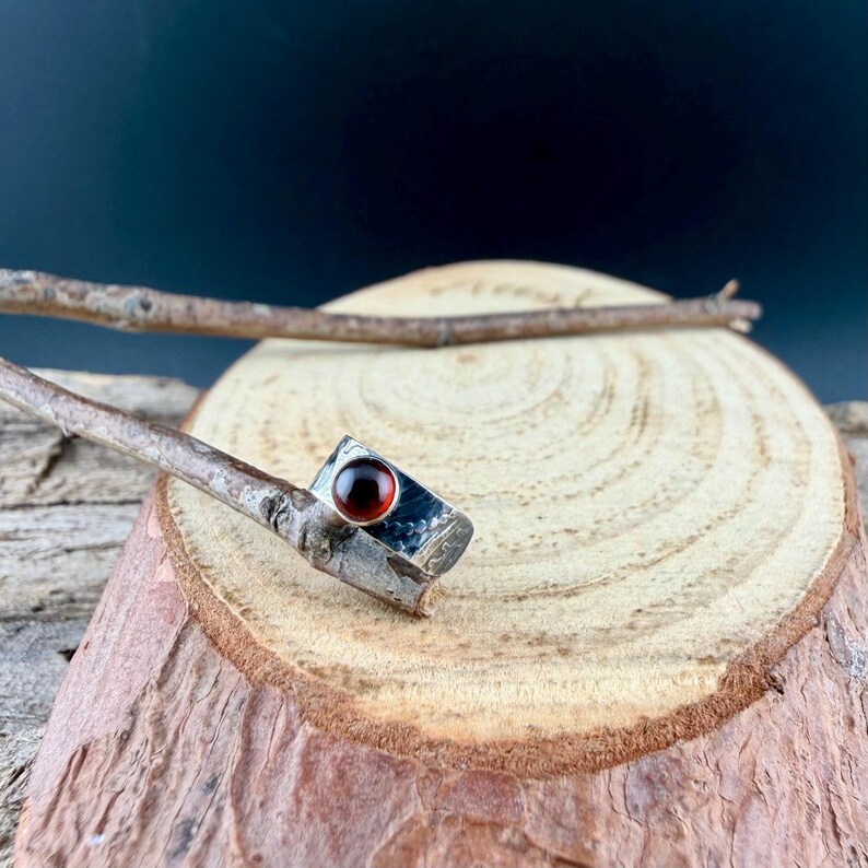 May include: A silver ring with a rectangular setting and a round, deep red gemstone. The ring is set against a natural wood background, with a dark backdrop. The ring's band has a textured appearance.