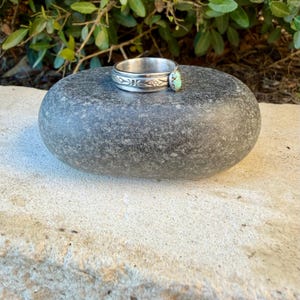 Puede incluir: Un anillo de plata con una banda que presenta patrones florales grabados y una peque&ntilde;a piedra turquesa de forma ovalada con vetas de matriz marr&oacute;n engastada en el lateral. La banda de metal tiene un acabado pulido y ancho uniforme.