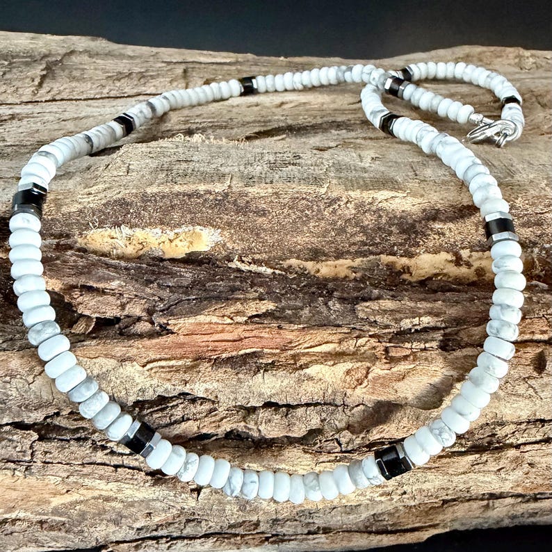 May include: White and gray beaded necklace with black accents. The necklace is composed of small, round, white beads interspersed with gray and black cylindrical beads. A silver clasp secures the necklace. The necklace is displayed on a wooden surface.