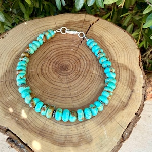 May include: A beaded necklace featuring round, turquoise-colored stones with brown matrix patterns. The beads are strung together with small silver-toned spacers and secured with a silver-toned lobster claw clasp at the top of the circular arrangement.