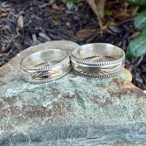 May include: Two silver-toned metal rings featuring a polished finish and a central band decorated with engraved geometric patterns. Each ring is bordered by a beaded edge detail, showcasing a consistent metallic texture and reflective surface.