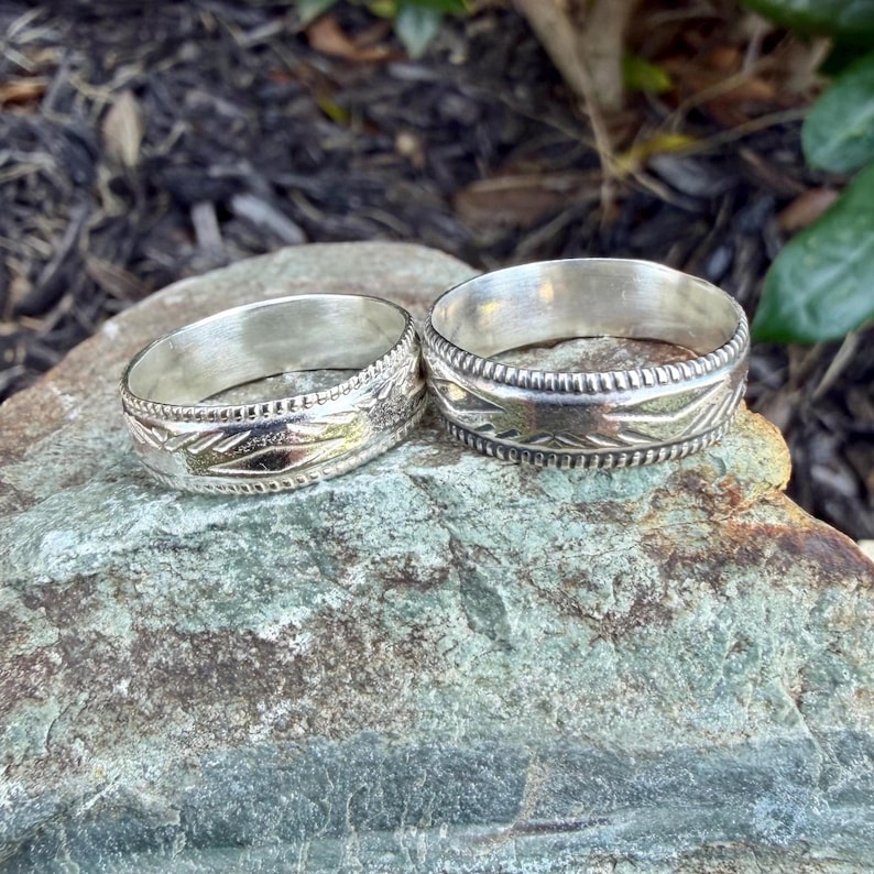 May include: Two silver-toned metal rings featuring a polished finish and a central band decorated with engraved geometric patterns. Each ring is bordered by a beaded edge detail, showcasing a consistent metallic texture and reflective surface.