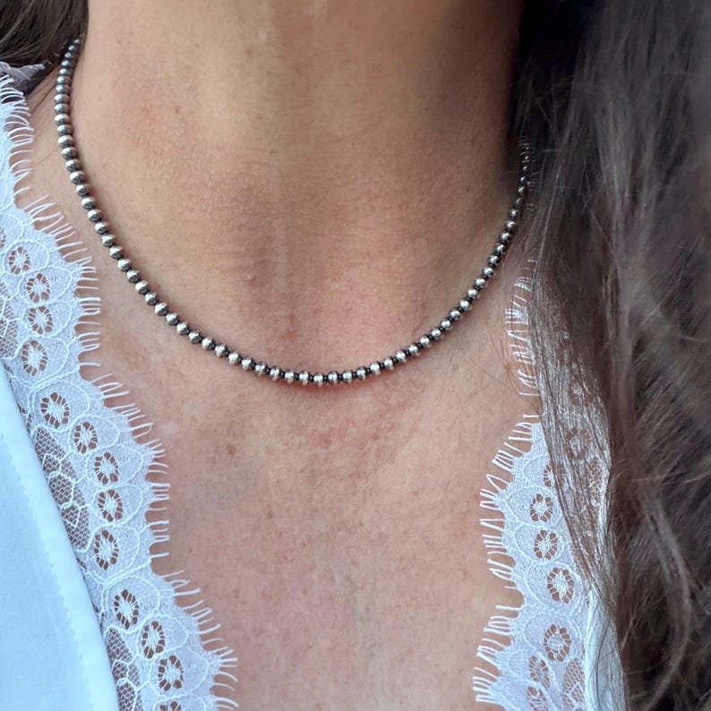 May include: A silver beaded necklace resting on a person's neck. The necklace is composed of small, round, silver beads. The person is wearing a white top with a lace trim.