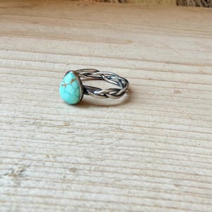 Puede incluir: Un anillo con una piedra turquesa en forma de l&aacute;grima con vetas marrones, engastada en una banda de metal oscuro con un dise&ntilde;o trenzado y retorcido. La piedra est&aacute; situada en la parte superior de la banda, que tiene un acabado oxidado y texturizado.